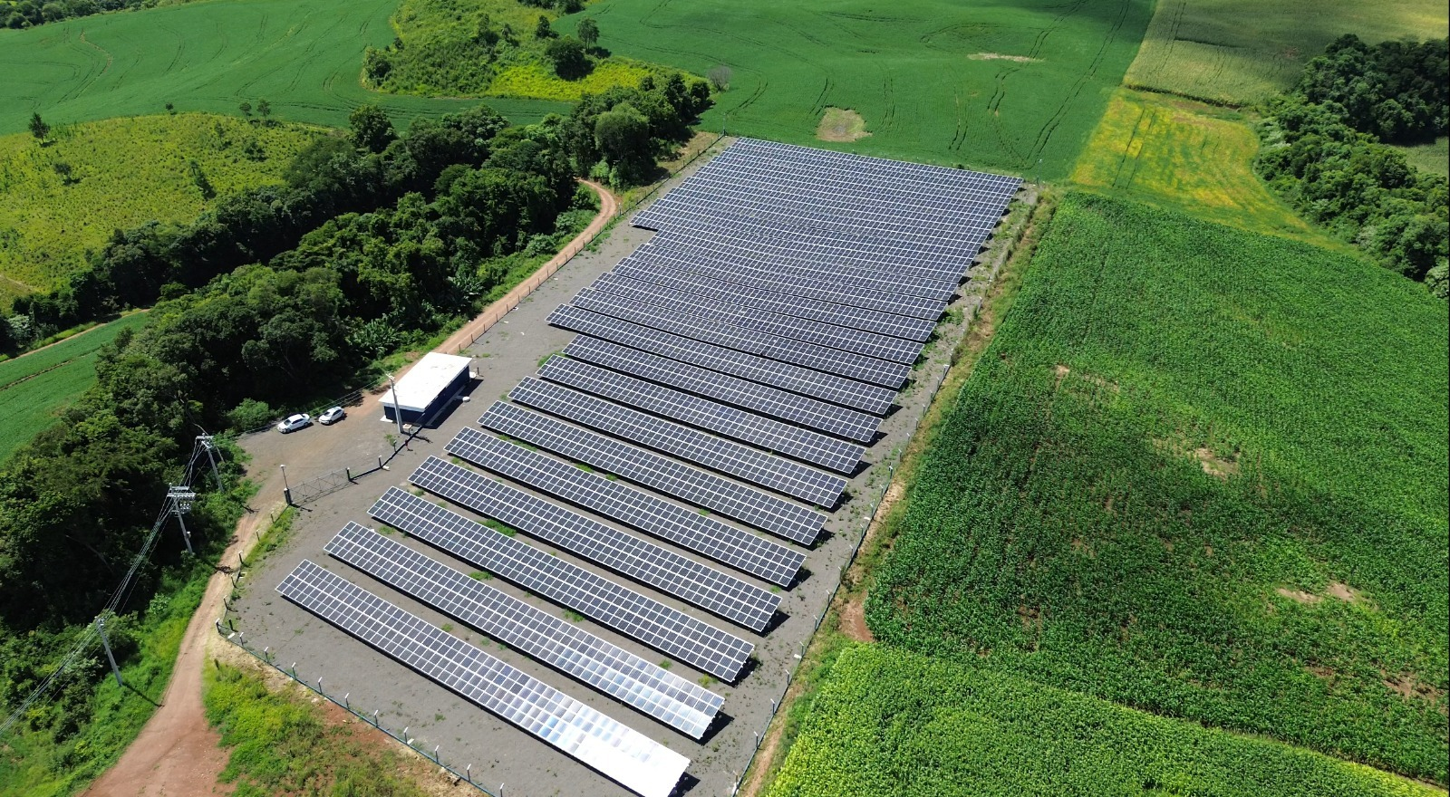 Aerial view of a large solar farm with multiple rows of photovoltaic panels installed on flat land, surrounded by lush green fields and trees. A small building and vehicles are visible near the entrance.